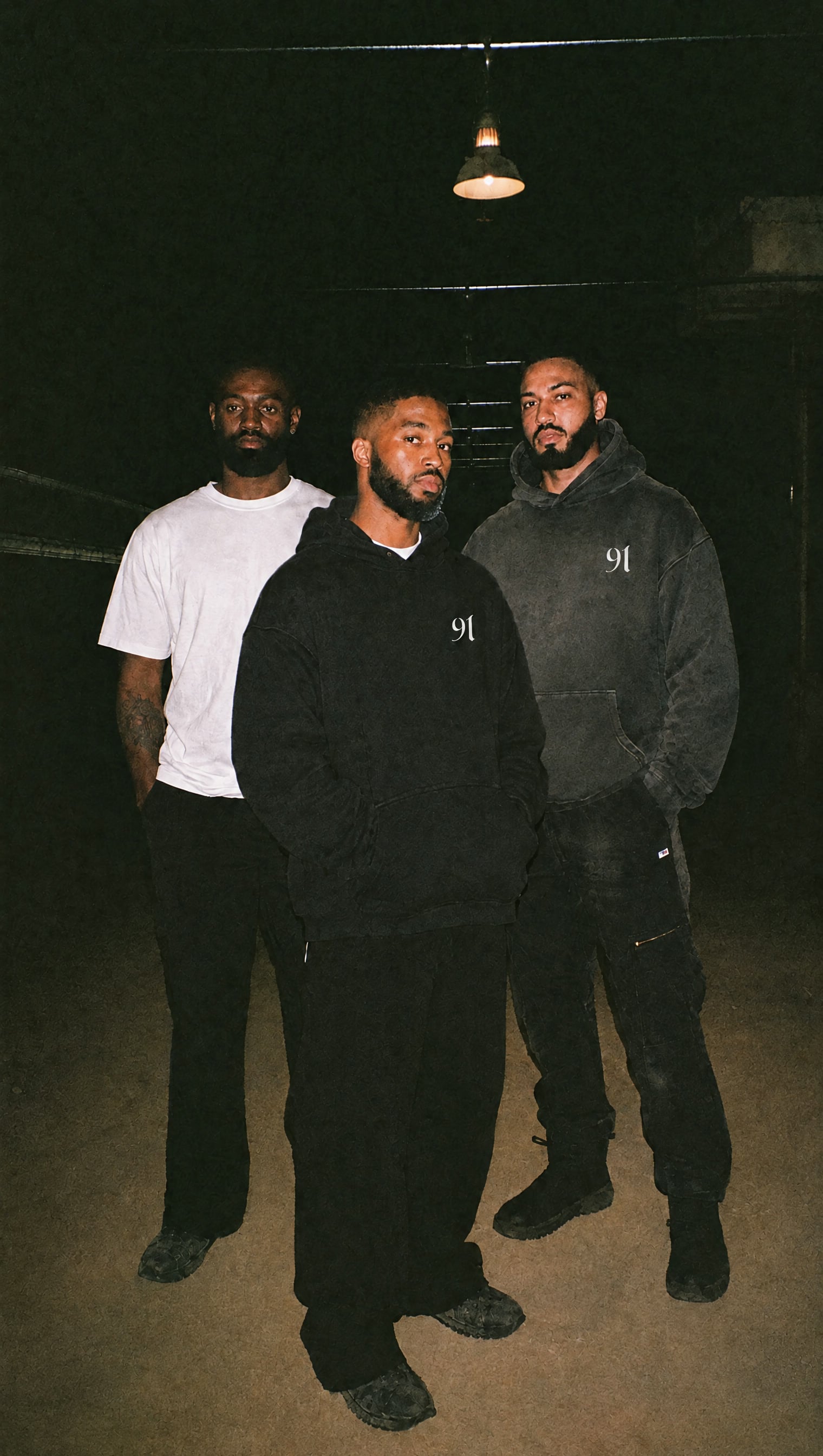 Two black heavyweight hoodies with the white 91 mark on the left chest, beside a plain white tee, under a single work lamp