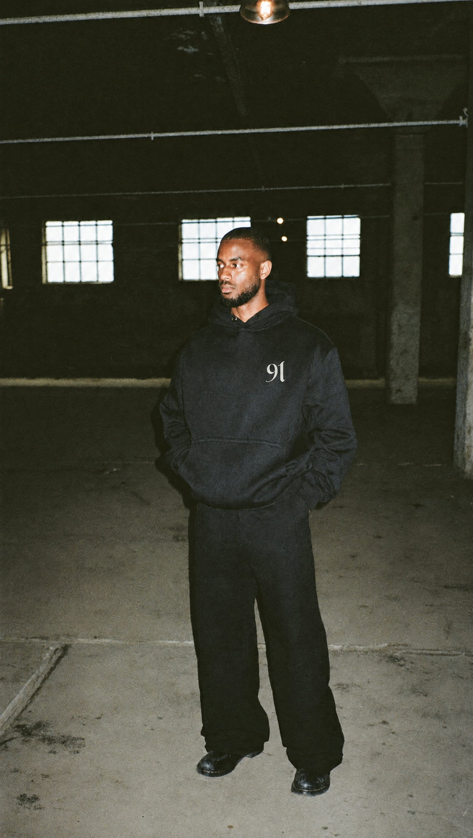 Black heavyweight hoodie with the small white 91 mark on the left chest, in a dark warehouse